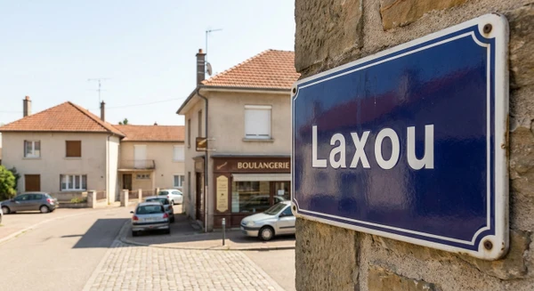Plan rapproché d'une plaque de rue ou d'une signalétique urbaine indiquant "Laxou" dans un cadre de petite commune en agglomération, avec en arrière-plan flou un environnement urbain typique de la banlieue nancéienne (petits commerces, rue calme, bâtiments résidentiels). Photo de jour en conditions d'éclairage naturel, perspective réaliste et professionnelle.